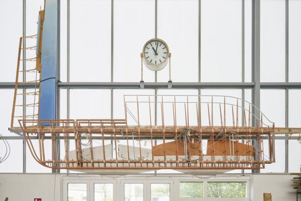 Das unfertige Holzmodell eines Bootes hängt in einer Werkshalle unter einer Uhr.