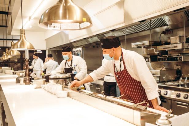 Mehrere Köche mit Masken bereiten Speisen in einer Restaurantküche zu.