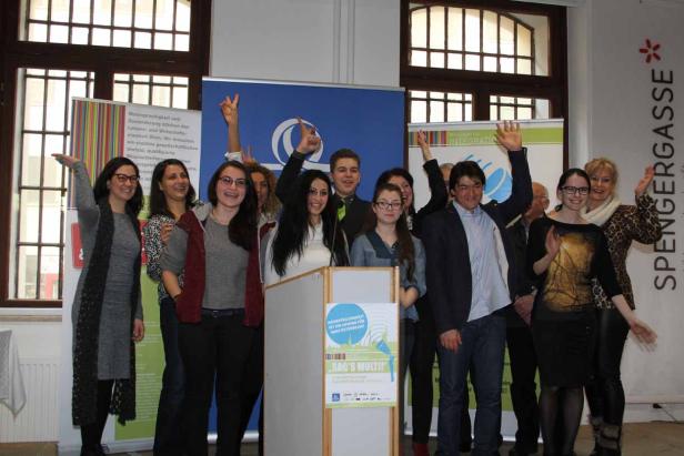 Eine Gruppe von Menschen steht hinter einem Rednerpult und winkt in die Kamera.