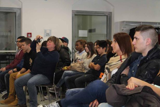 Eine Gruppe von Menschen sitzt auf Stühlen und schaut in eine Richtung. Eine Frau macht Fotos.