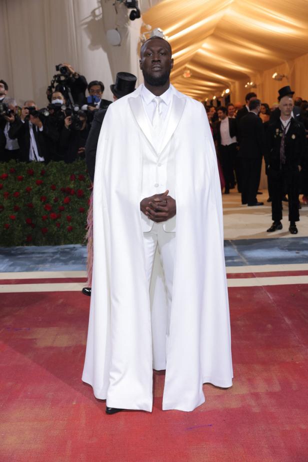 Omar Sy in einem weißen Anzug und Umhang auf dem roten Teppich.