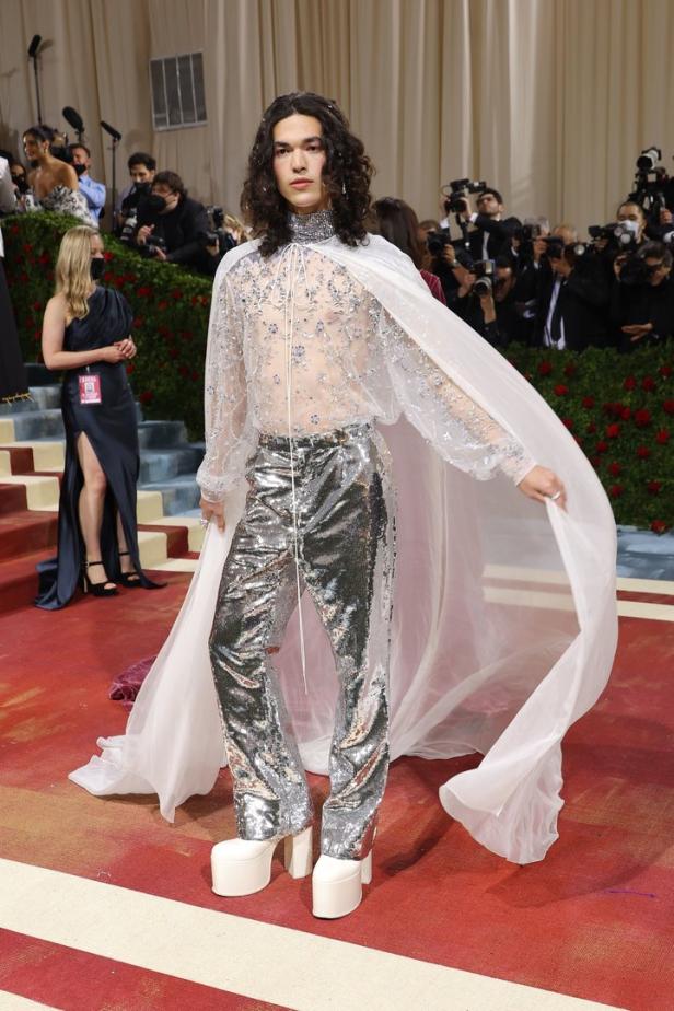 Ein Mann mit langem, lockigem Haar trägt ein glitzerndes Outfit und einen weißen Umhang auf dem roten Teppich.