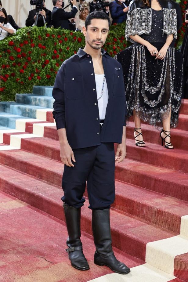 Riz Ahmed auf dem roten Teppich in einem marineblauen Outfit und Stiefeln.