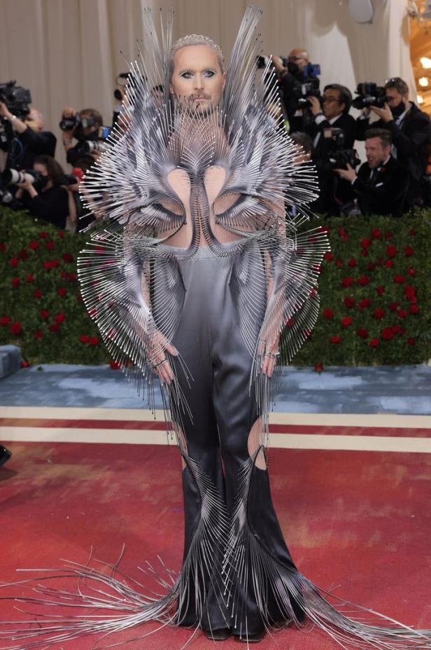 Ein Mann in einem extravaganten, silberfarbenen Outfit posiert auf einem roten Teppich.