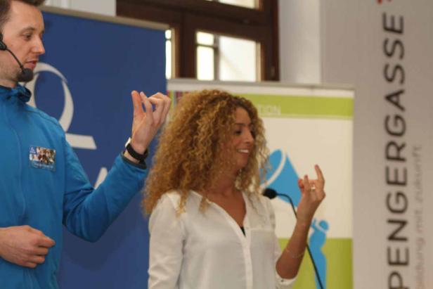 Eine Frau mit lockigem Haar gestikuliert während einer Präsentation neben einem Mann mit Headset.