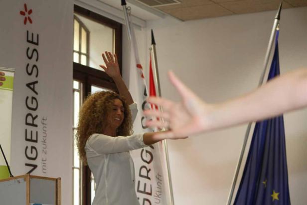 Eine Frau mit lockigem Haar gestikuliert vor Bannern und einer Europaflagge.