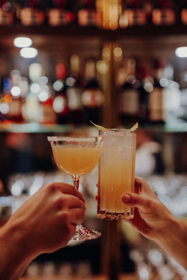 Zwei Personen stoßen mit Cocktails vor einer Bar an.