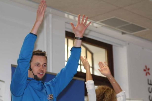 Ein Mann mit Mikrofon gestikuliert mit erhobenen Armen vor einer Gruppe von Menschen.