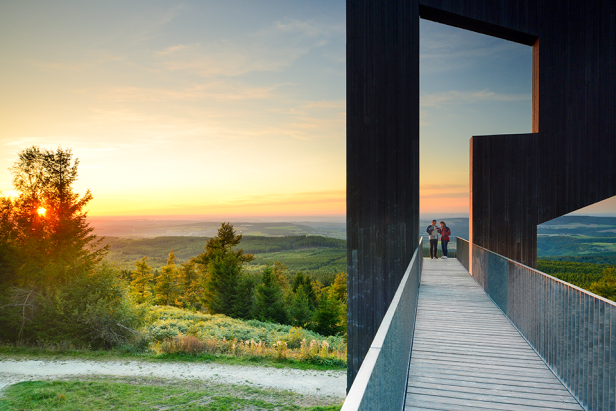 Zwei Personen auf einer Aussichtsplattform mit Blick auf eine bewaldete Landschaft bei Sonnenuntergang.