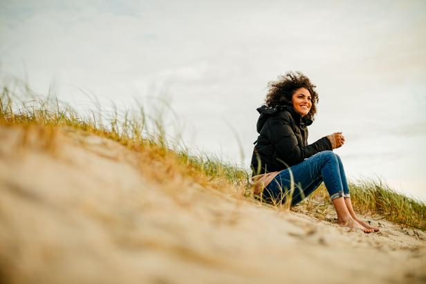 Eine Frau sitzt barfuß in Jeans und Jacke auf einer Sanddüne.