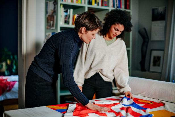Zwei Frauen betrachten einen Stoff mit einem rot-weiß-blauen Muster auf einem Tisch.