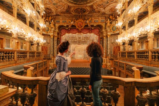 Zwei Frauen blicken von einem Balkon auf die Bühne eines opulenten Theaters.