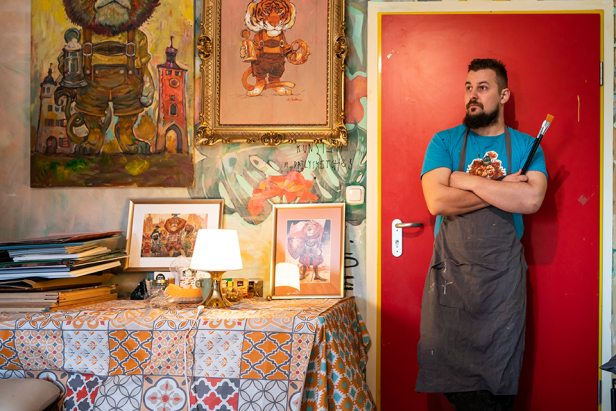 Ein bärtiger Mann mit verschränkten Armen und Pinsel steht vor einer roten Tür in seinem Atelier.