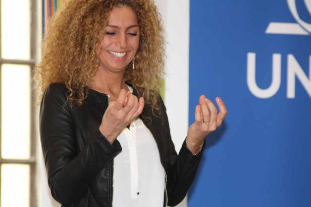 Eine Frau mit lockigem Haar lächelt und gestikuliert vor einem blauen Hintergrund mit dem Aufdruck „UN“.