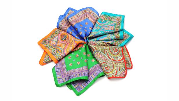 Mehrere farbenfrohe Einstecktücher mit verschiedenen Mustern liegen kreisförmig auf einem weißen Hintergrund.