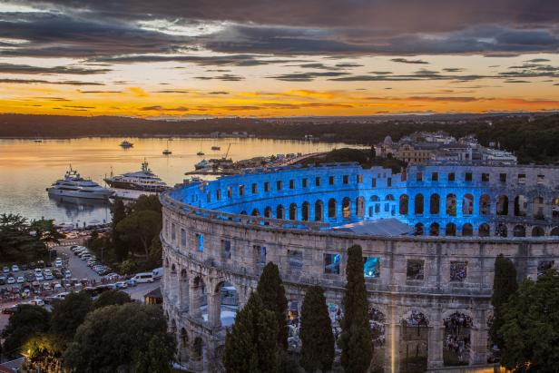 Das Amphitheater von Pula, beleuchtet in blau, mit Yachten im Hintergrund bei Sonnenuntergang.