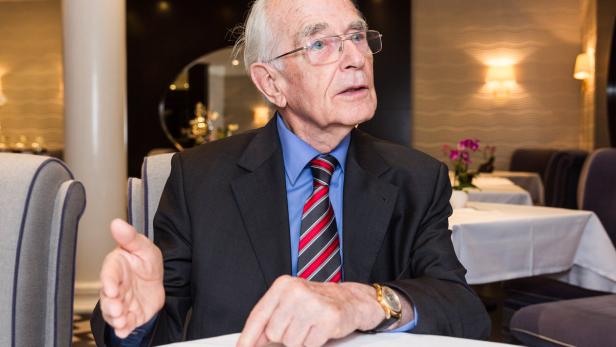 Ein älterer Mann mit Brille und Anzug sitzt an einem Tisch in einem Restaurant.