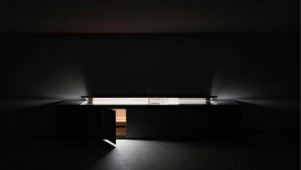 Ein dunkler Raum mit einer beleuchteten Theke und einer teilweise geöffneten Tür, die einen Blick auf eine Sauna freigibt.