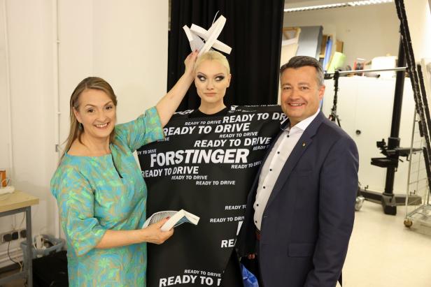 Eine Frau mit Papierflieger-Kopfschmuck posiert mit zwei Personen vor einem Banner von Forstinger „Ready to Drive“.