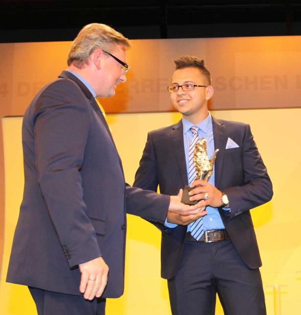 Ein Mann überreicht einem jüngeren Mann eine goldene Auszeichnung auf einer Bühne.