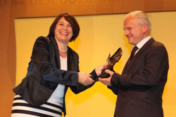 Eine lachende Frau nimmt eine goldene Adler-Statue von einem Mann entgegen.