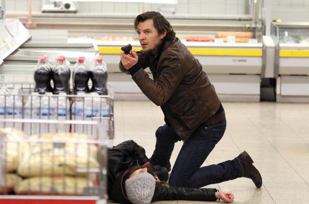 Ein Mann mit einer Waffe kniet neben einer am Boden liegenden Frau in einem Supermarkt.