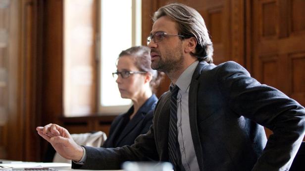 Ein Mann mit Brille gestikuliert während einer Besprechung, im Hintergrund eine Frau.