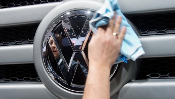 Eine Hand poliert mit einem blauen Tuch das VW-Logo an einem grauen Auto.