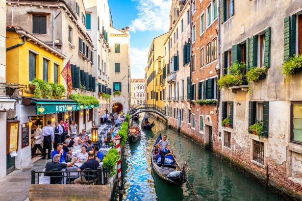 Eine belebte Szene in Venedig mit Gondeln auf einem Kanal und Gästen in einem Restaurant.