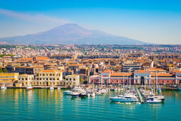 Blick auf Catania mit dem Ätna im Hintergrund und einem Hafen voller Boote im Vordergrund.