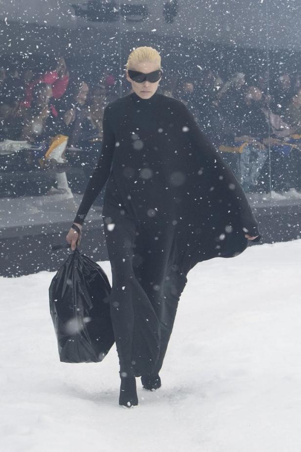 Eine Frau in einem schwarzen Kleid und futuristischer Brille trägt eine Mülltüte im Schnee.