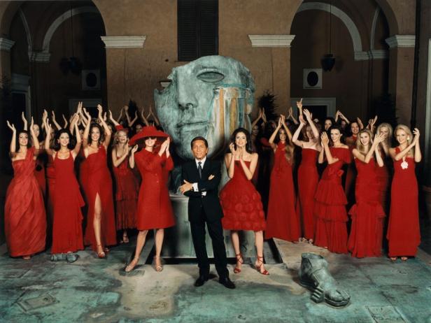 Valentino steht vor einer Gruppe von Frauen in roten Kleidern und einer großen Skulptur.