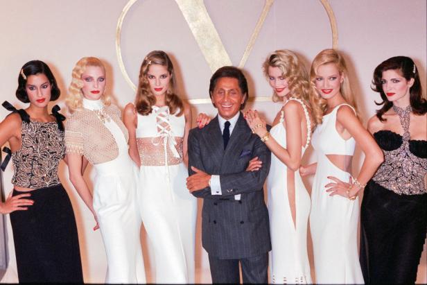 Valentino posiert mit sechs Models in weißer und schwarzer Abendgarderobe vor einem goldenen „V“.