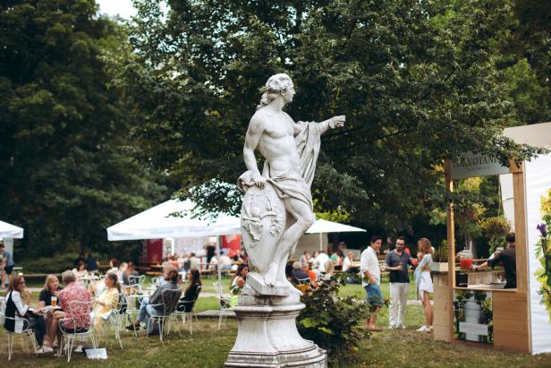 Eine Statue steht in einem Park, während sich Menschen an Tischen und einer Bar versammeln.