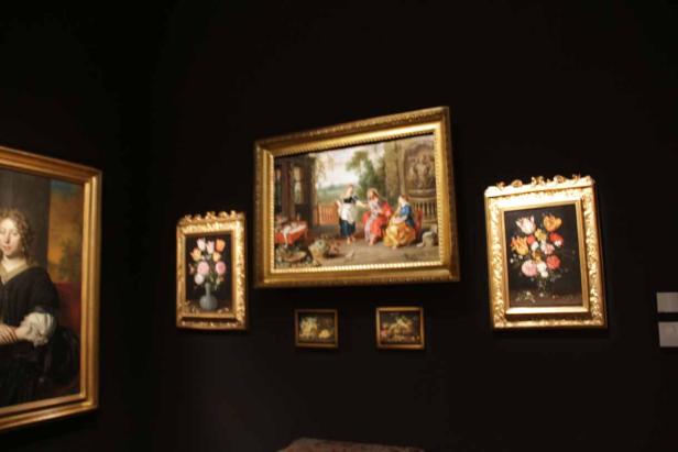 Eine Auswahl an Gemälden mit goldenen Rahmen hängt an einer dunklen Wand.