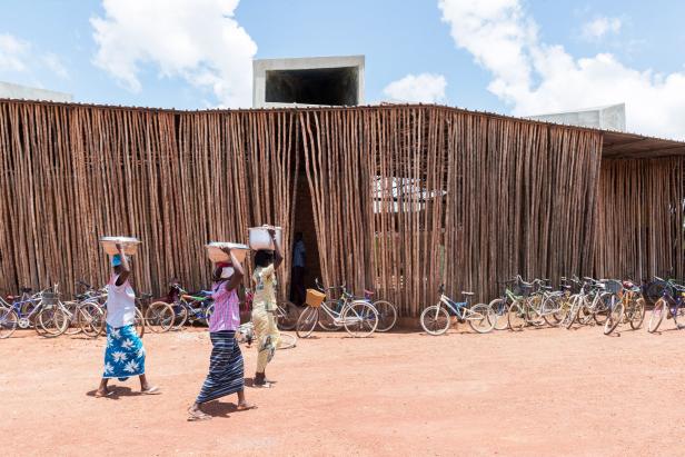 Drei Frauen tragen Schüsseln auf dem Kopf vor einem Gebäude mit einer Fassade aus Holzstämmen.