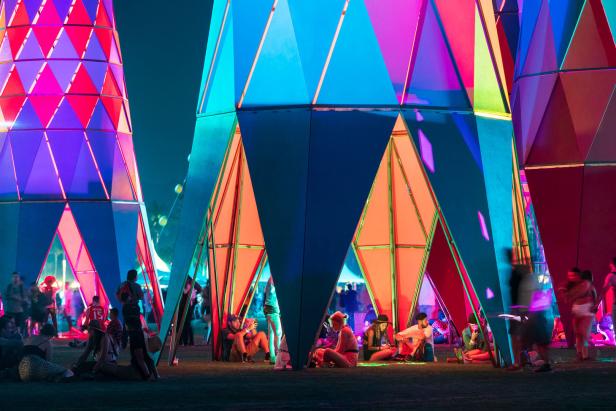 Menschen sitzen und stehen unter farbenfrohen, geometrischen Installationen bei Nacht.