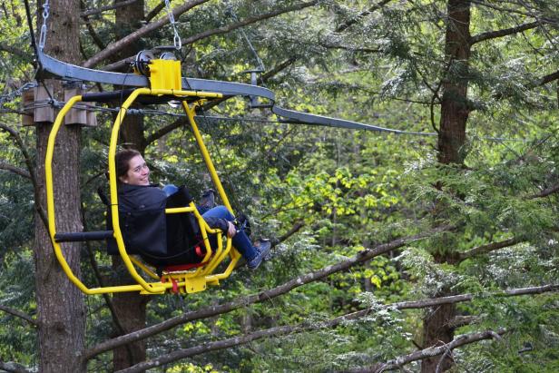 Eine Frau fährt mit einem gelben Sessellift durch einen Wald.