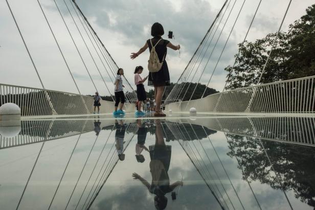Eine Frau macht ein Selfie auf einer Glasbrücke, während andere Besucher im Hintergrund zu sehen sind.