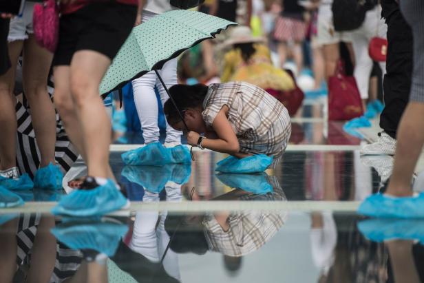 Besucher mit blauen Überschuhen auf einer Glasbrücke, eine Frau macht ein Foto.