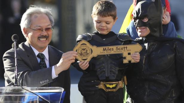 Ein Mann im Anzug, ein Kind im Batman-Kostüm und ein Mann als Batman halten einen goldenen Schlüssel für Miles Scott.