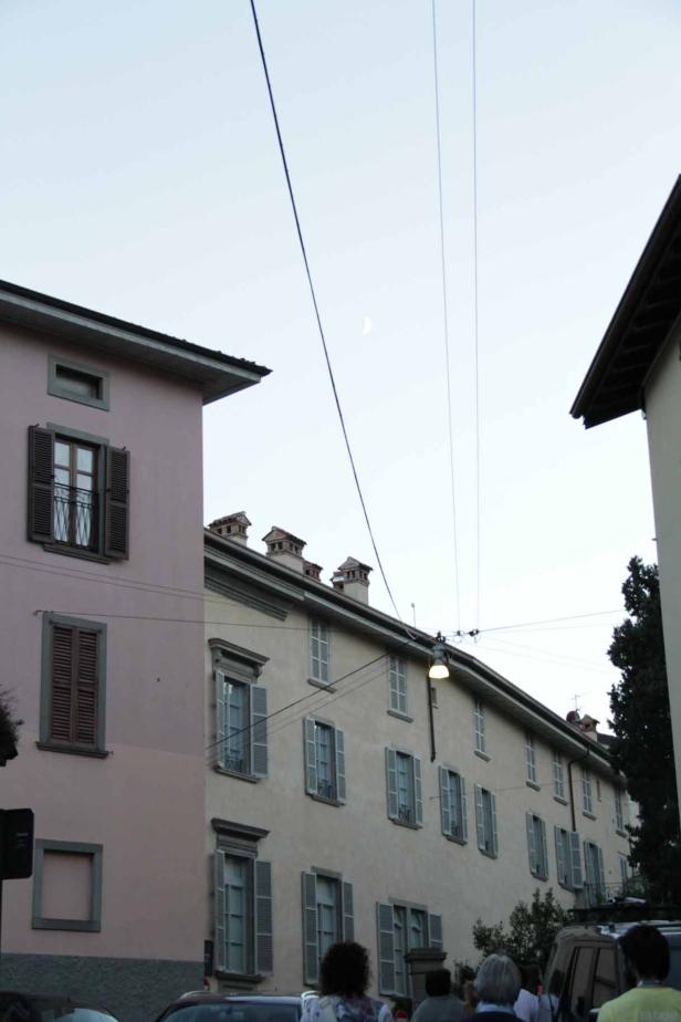 Blick zwischen Häusern hindurch auf eine schmale Mondsichel am Himmel.