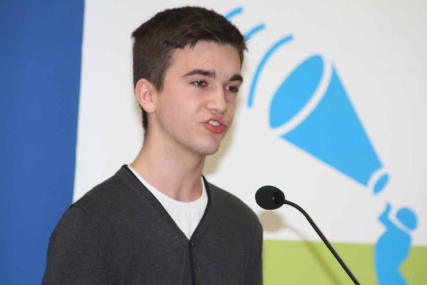 Ein junger Mann spricht am Mikrofon vor einem blauen Logo.