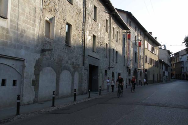 Zwei Personen fahren mit Fahrrädern eine Straße entlang, gesäumt von alten Steingebäuden.