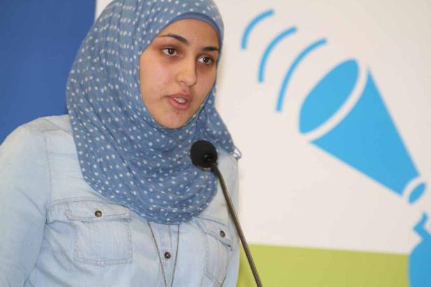 Eine Frau mit blauem Kopftuch spricht an einem Mikrofon.