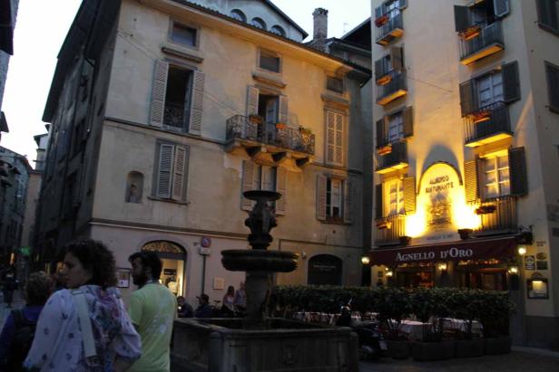 Ein Platz in einer italienischen Stadt mit einem Brunnen und dem Restaurant „Agnello D’Oro“.
