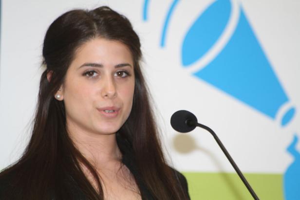 Eine junge Frau spricht an einem Mikrofon vor einem blauen Logo.