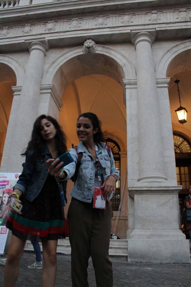 Zwei junge Frauen machen ein Selfie vor einem Gebäude mit Säulen und Bögen.