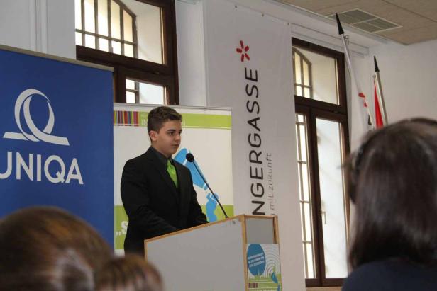 Ein junger Mann hält eine Rede an einem Rednerpult vor Bannern von UNIQA und der NMS Rengersgasse.