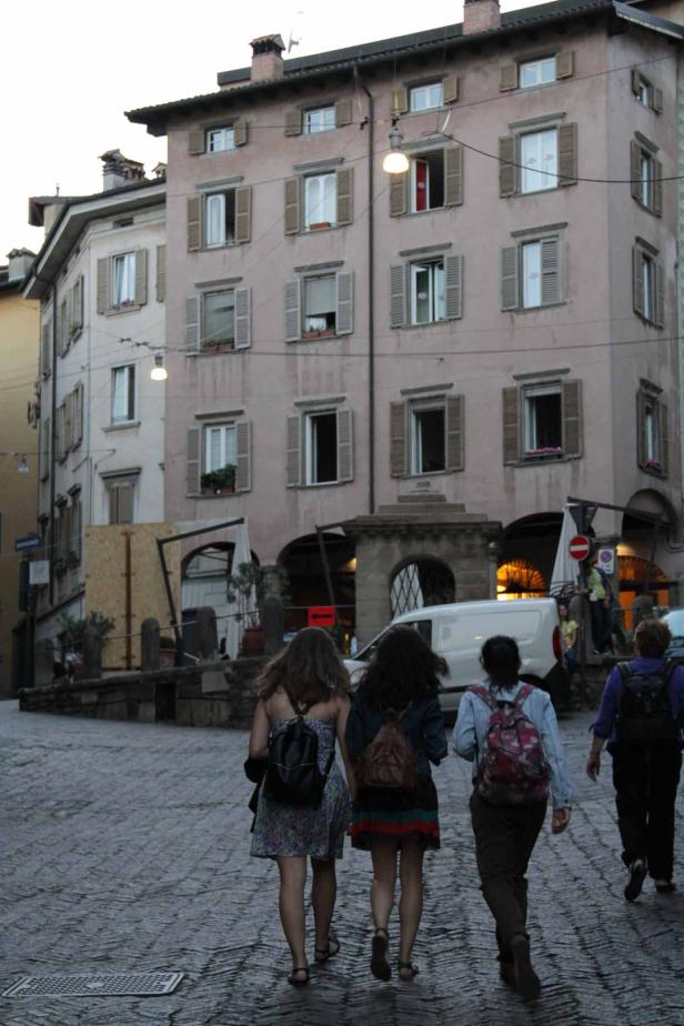 Vier Personen gehen eine Kopfsteinpflasterstraße entlang, im Hintergrund ein mehrstöckiges Gebäude.